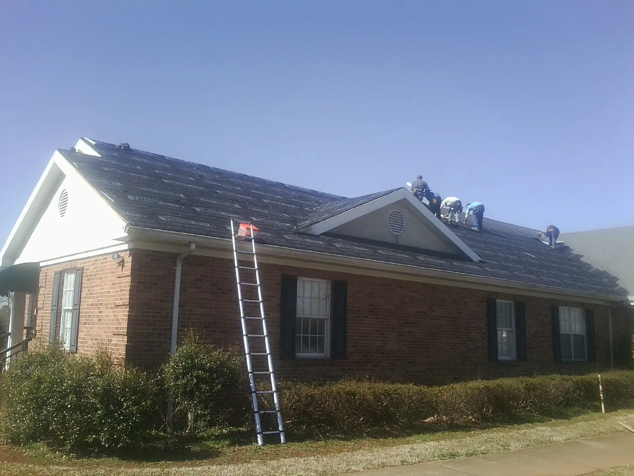 Crew tearing shingles off a brick ranch roof with a ladder set — Emergency Roof Repair work by our crew in North Grafton