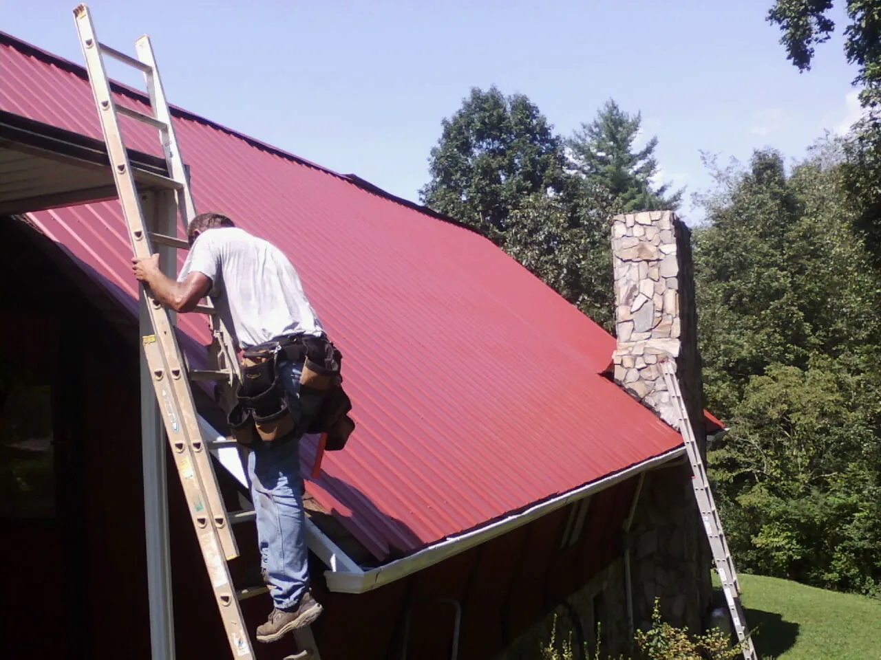 Roofer on a ladder beside a red metal roof and stone chimney — Soffit & Fascia Repair by Roofing Brothers in North Grafton